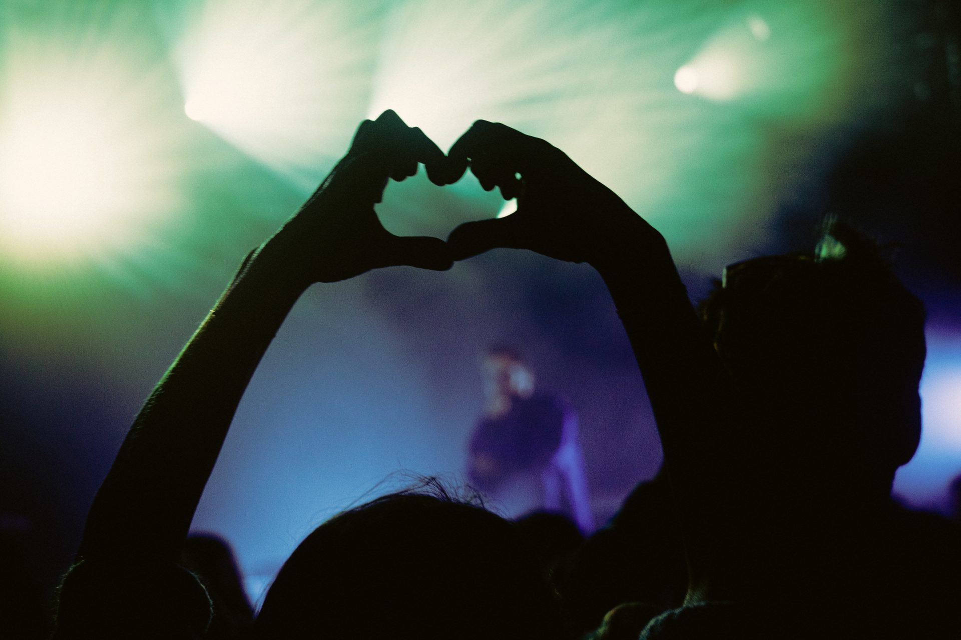 Une personne forme un coeur avec ses doigts au premier plan. En arrière plan, l'artiste Achile