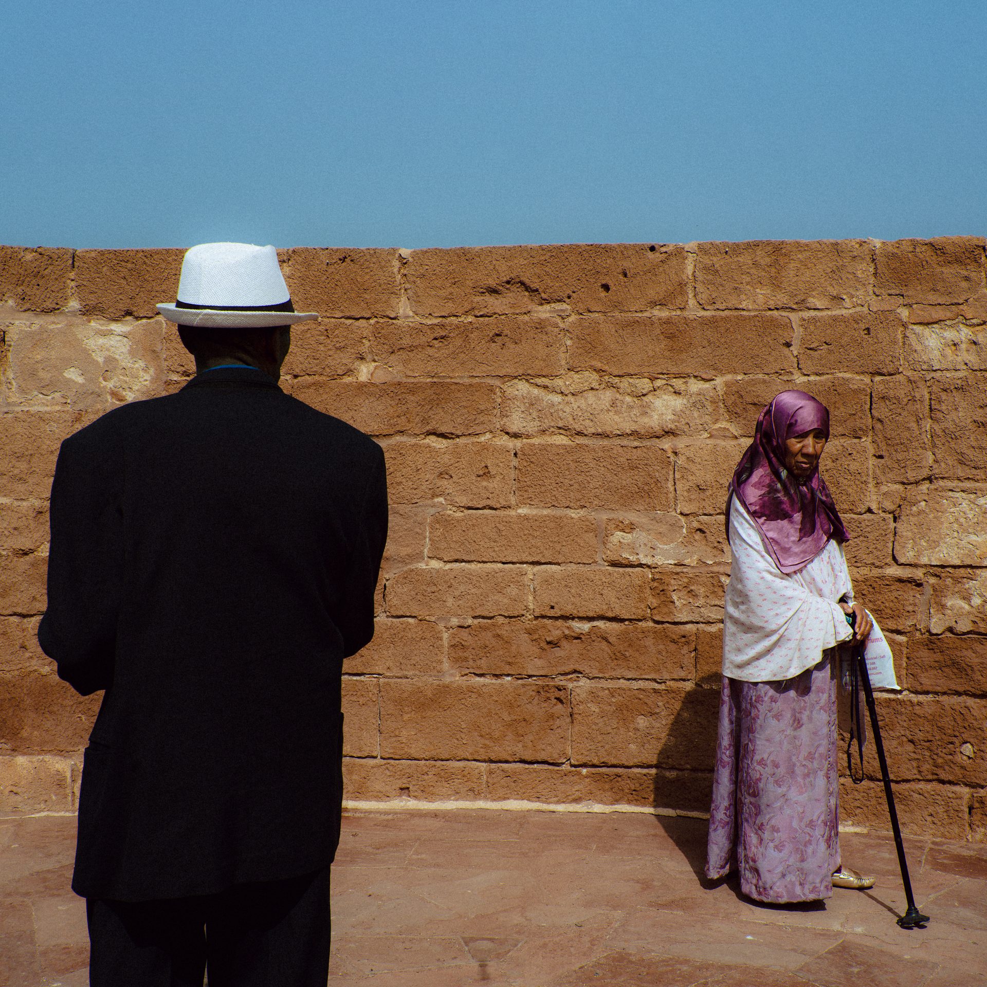 Un homme en costume noir et avec un chapeau blanc regarde sa femme vétue d'un voile et d'une robe rose. Les deux font face à un mur d'Essaouira.
