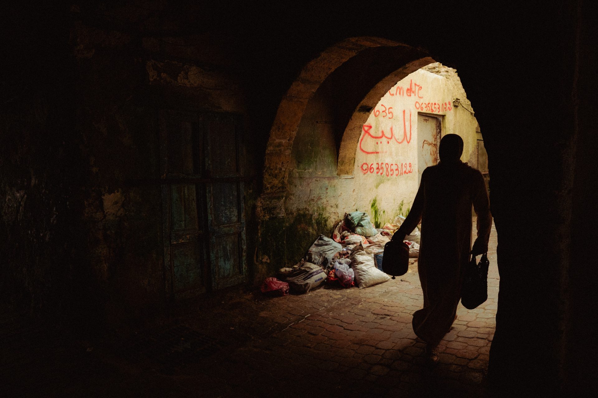 Une femme tenant des sacs passe sous un pont. Dans le fond, la lueur du jour illumine des dêchets.